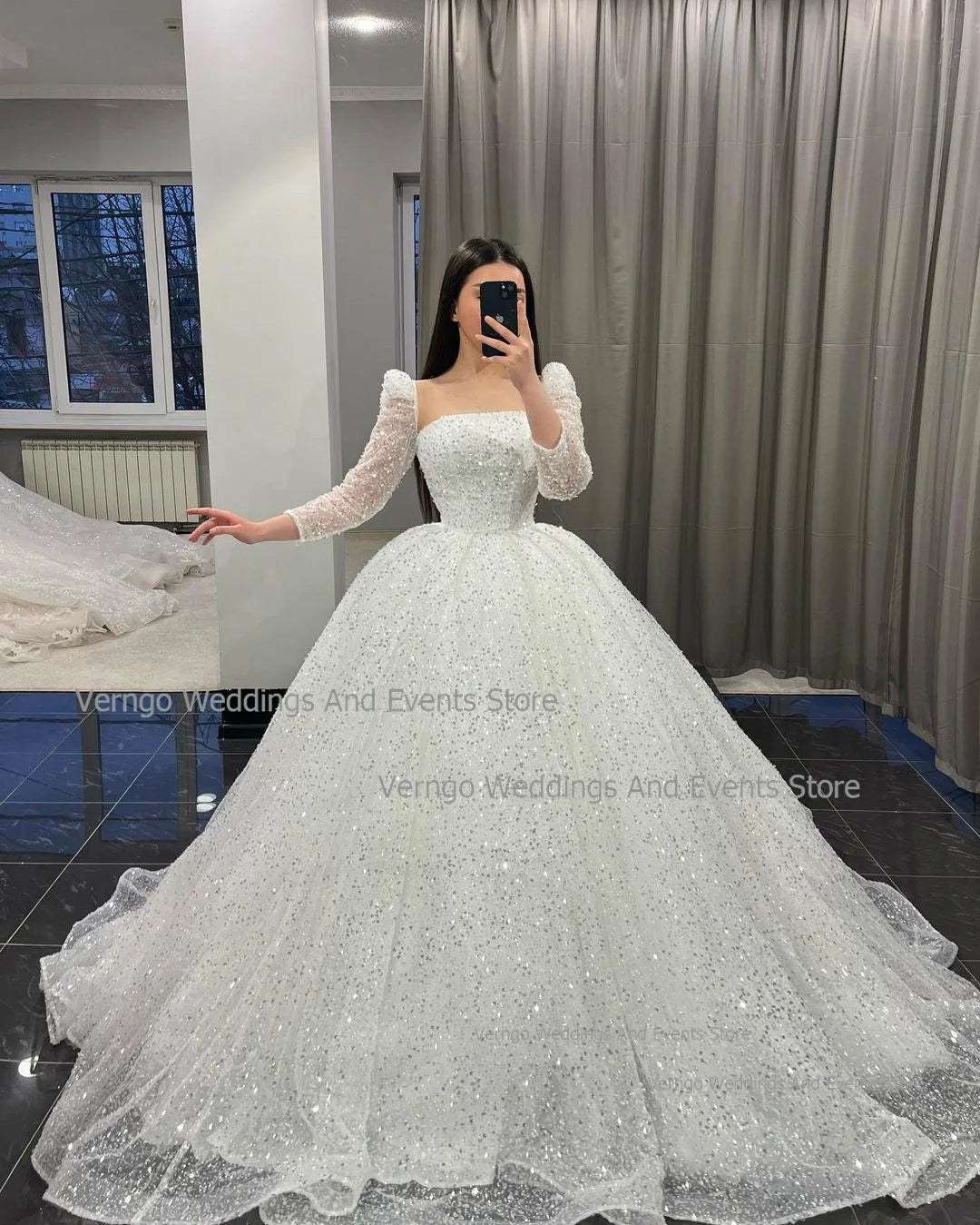 A woman in a sparkling white wedding gown with long lace sleeves takes a mirror selfie in a room with gray curtains and a window showing trees outside.