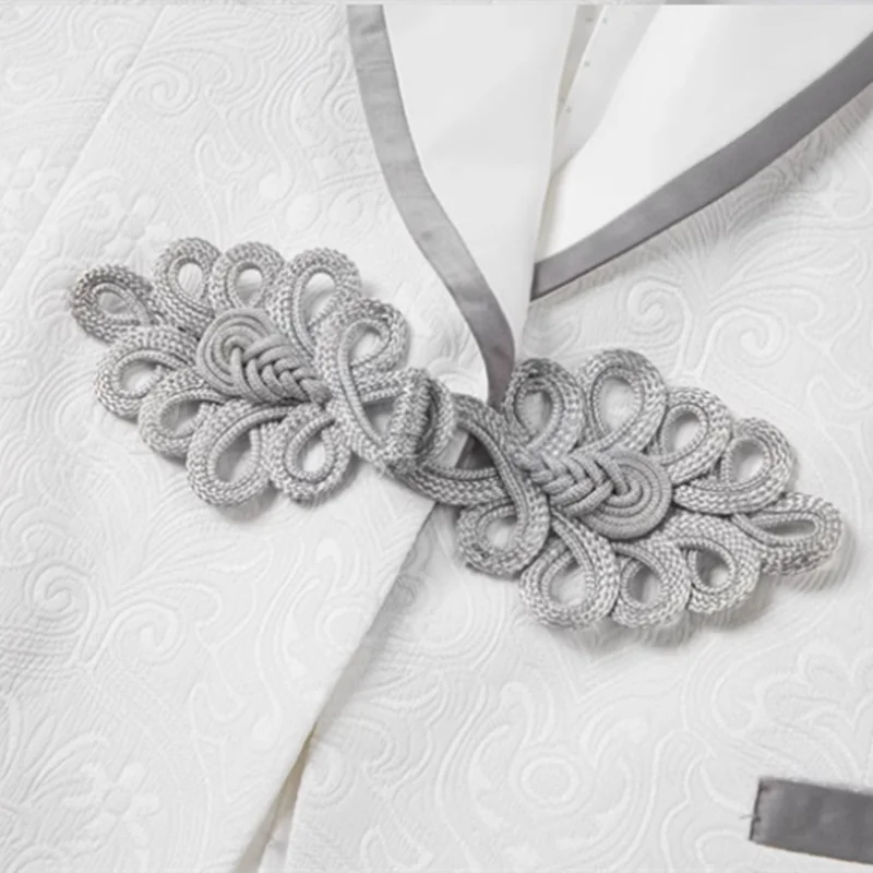Close-up of an ornate silver knot embellishment on a textured white garment with a gray trim. The fabric features an intricate floral pattern.