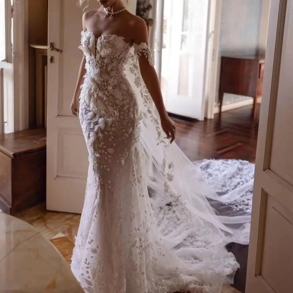 A bride in an elegant, off-the-shoulder lace wedding gown with a long train and floral appliques stands in a softly lit room with wooden floors and an open door.