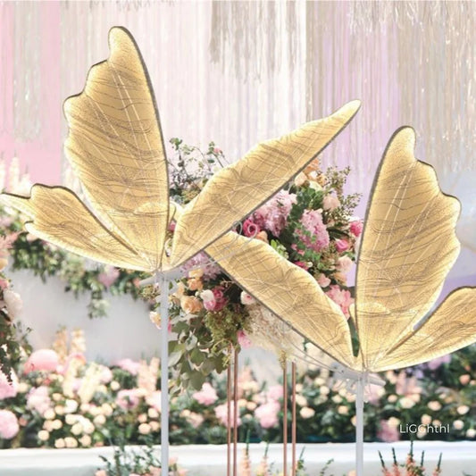 Ornate golden fairy wings stand amidst lush floral arrangements with pastel flowers, set against a backdrop of shimmering curtains, creating an ethereal atmosphere.
