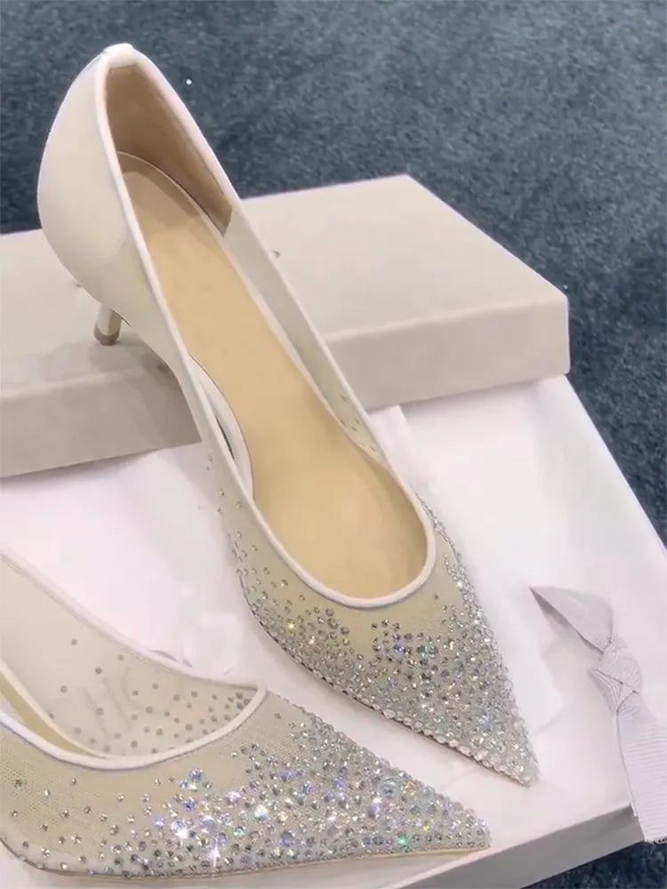 Elegant high-heeled shoes with pointed toes, adorned with sparkling rhinestones on a sheer fabric, set on a white box against a gray carpet backdrop.