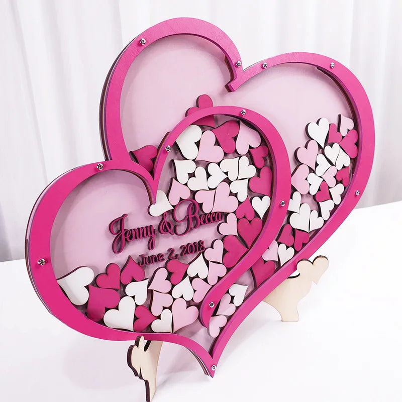 Pink and white heart-shaped guest book with small wooden hearts inside. Text reads "Jenny & Becca, June 2, 2018." The decoration stands on a light background.