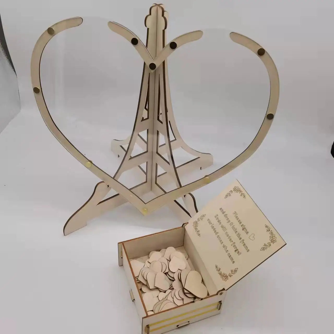 Wooden heart-shaped stand with an open box containing small heart cutouts. The box lid features decorative text and floral designs. Everything is made from light-colored wood.