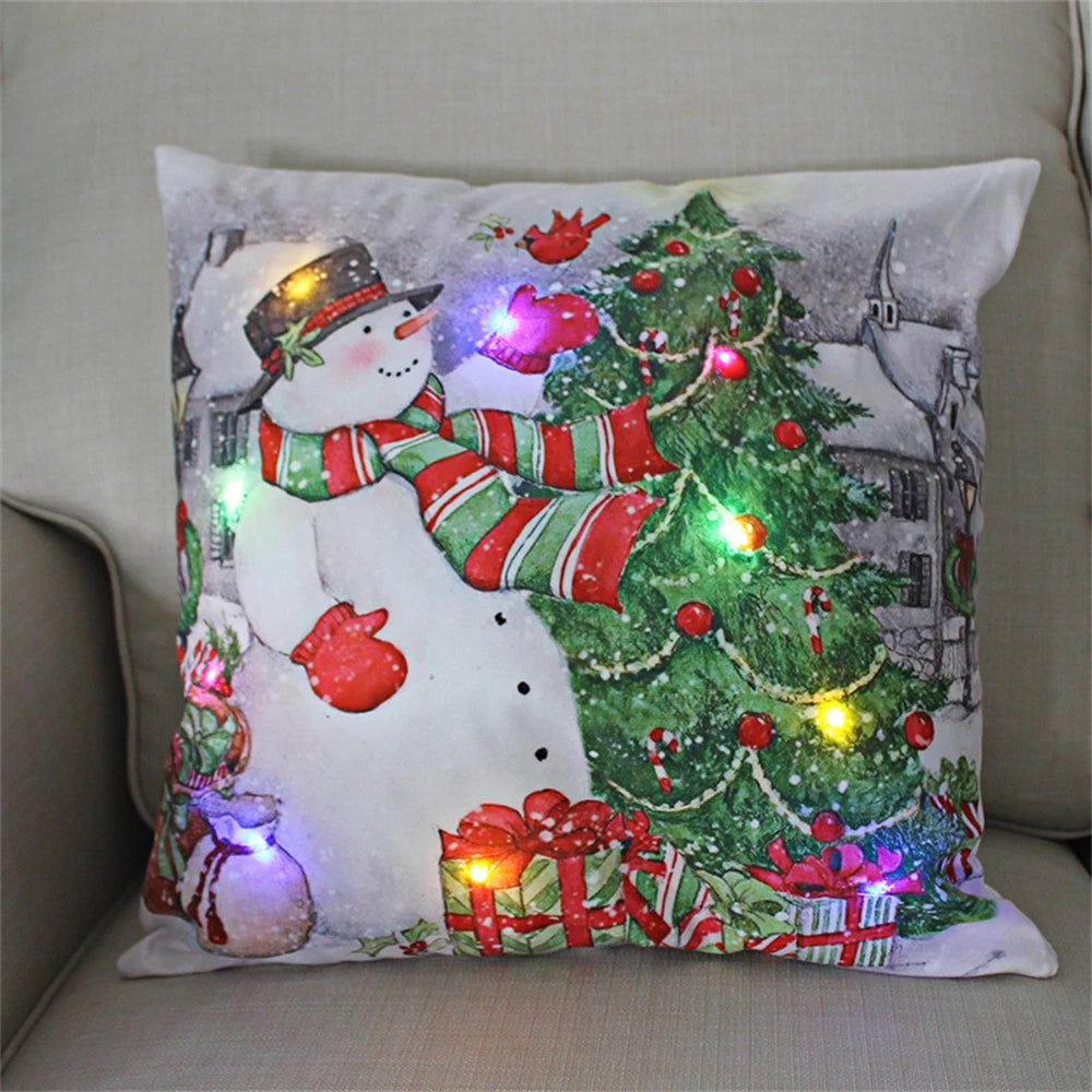Festive pillow on sofa featuring a snowman with a striped scarf, surrounded by gifts, a decorated Christmas tree, and twinkling colorful lights.