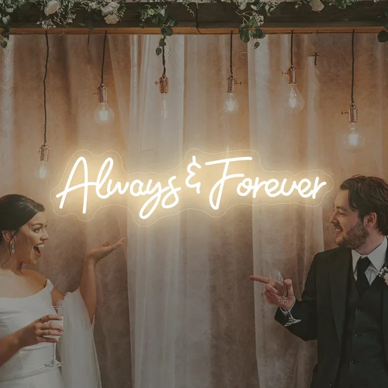 Bride and groom smiling and pointing at a glowing neon sign that reads "Always & Forever," surrounded by hanging lights and flowers, creating a romantic atmosphere.