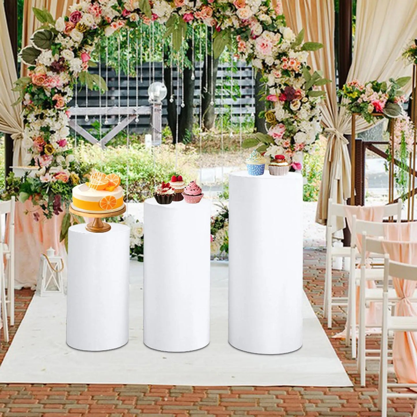 Wedding scene with floral arch and white columns. A cake with orange slices and various cupcakes are displayed on the columns. Rows of white chairs flank a brick pathway.