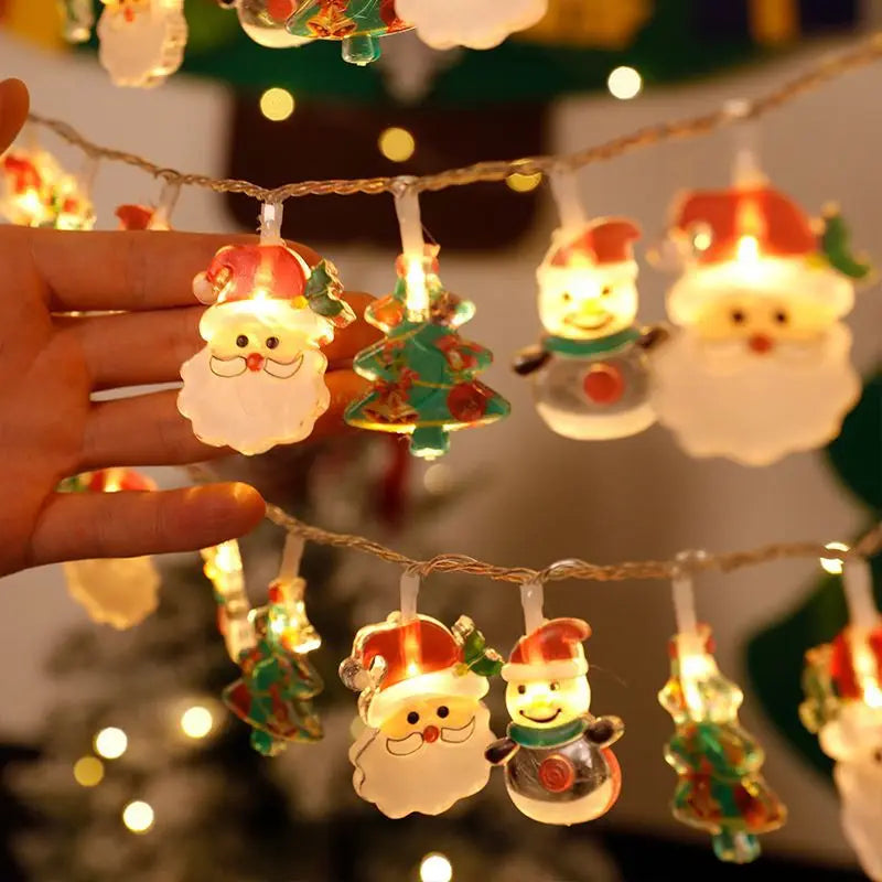 A hand adjusts glowing Christmas lights shaped like Santa, a snowman, and Christmas trees. The warm light creates a festive and cozy atmosphere.