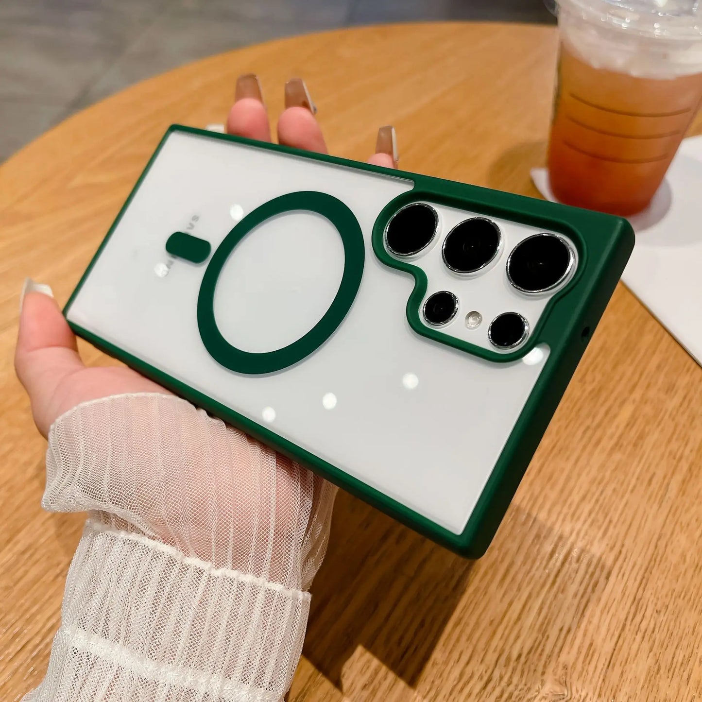 A person holds a smartphone with a stylish case featuring a green border, circular accents, and multiple camera lenses. A drink sits on a wooden table in the background.