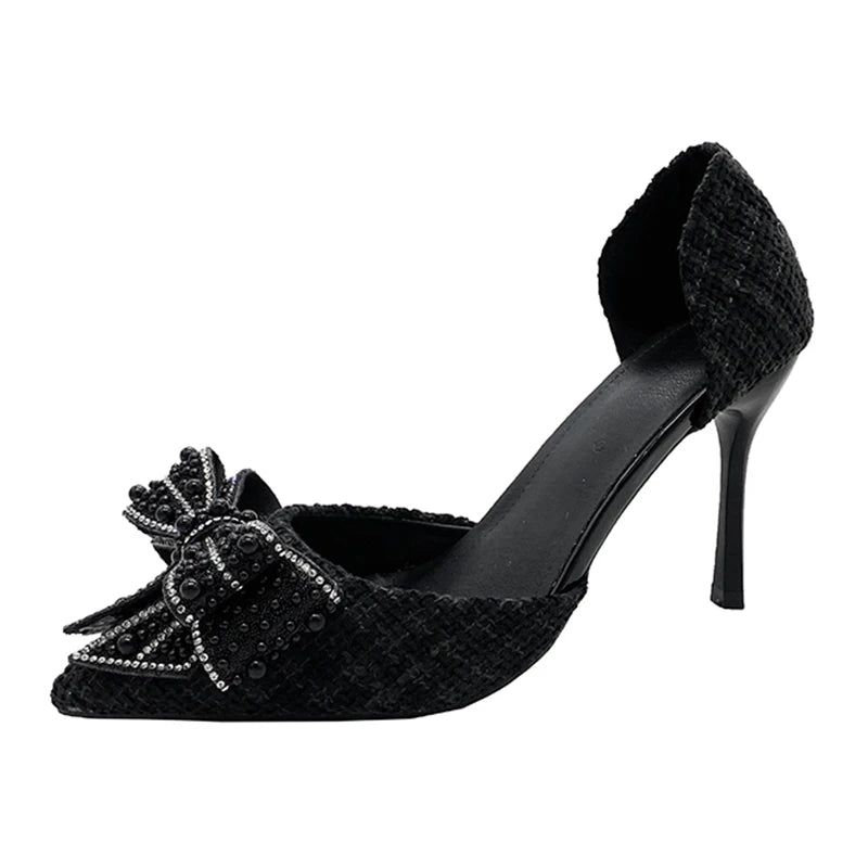 Black high-heeled shoe with a pointed toe and a decorative beaded bow. The heel is slender, and the shoe has a textured fabric, creating an elegant and sophisticated appearance.