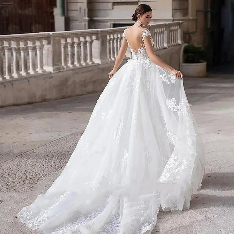 A bride in a flowing white lace wedding gown with an open back stands outdoors on a stone pathway, holding the hem of her dress, near an elegant stone balustrade.