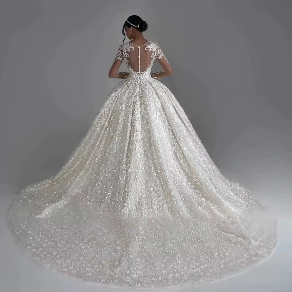 A bride in an elegant gown with intricate lace and a long train stands with her back to the camera. Her hair is styled in a bun, adorned with a headpiece.