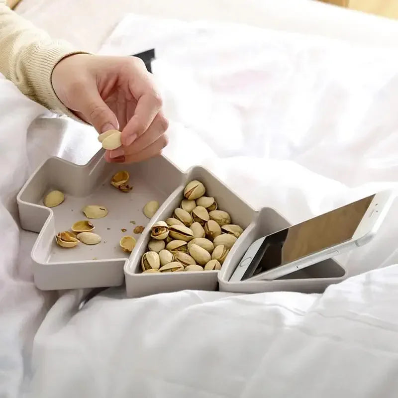Hand picking pistachios from a star-shaped tray with separate sections for nuts, shells, and a smartphone, set on a bed with white sheets.