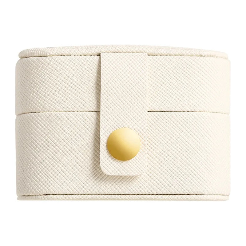 Cream-colored round jewelry box with a textured surface, secured by a strap featuring a gold button, against a white background.