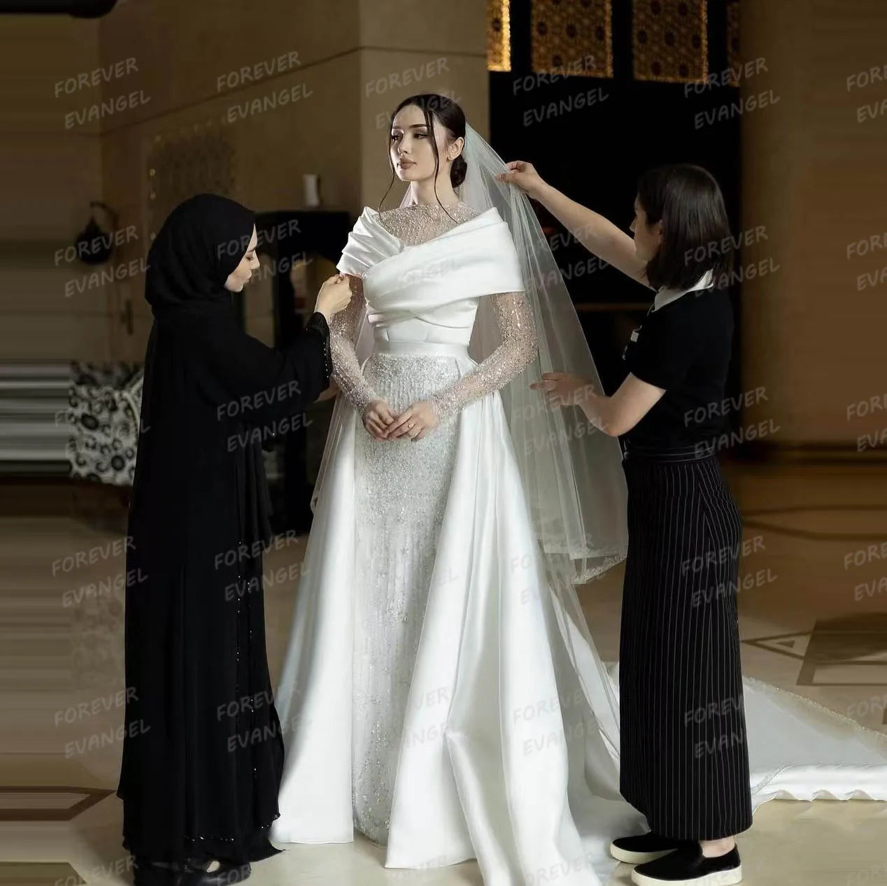 Bride wearing an elegant white gown with a long veil, attended by two assistants adjusting her attire in a warmly lit room. The dress features intricate lace and draped fabric.