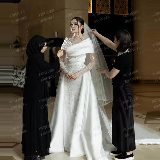 Bride wearing an elegant white gown with a long veil, attended by two assistants adjusting her attire in a warmly lit room. The dress features intricate lace and draped fabric.