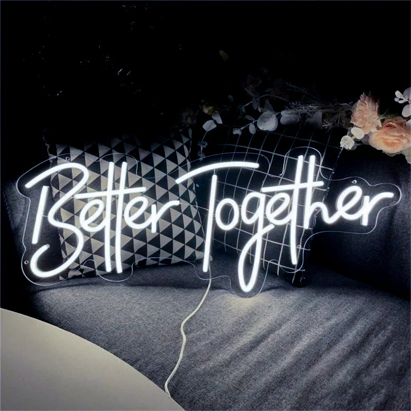 Neon sign reading "Better Together" in white cursive on a dark background, with decorative pillows and peach flowers nearby, creating a cozy ambiance.