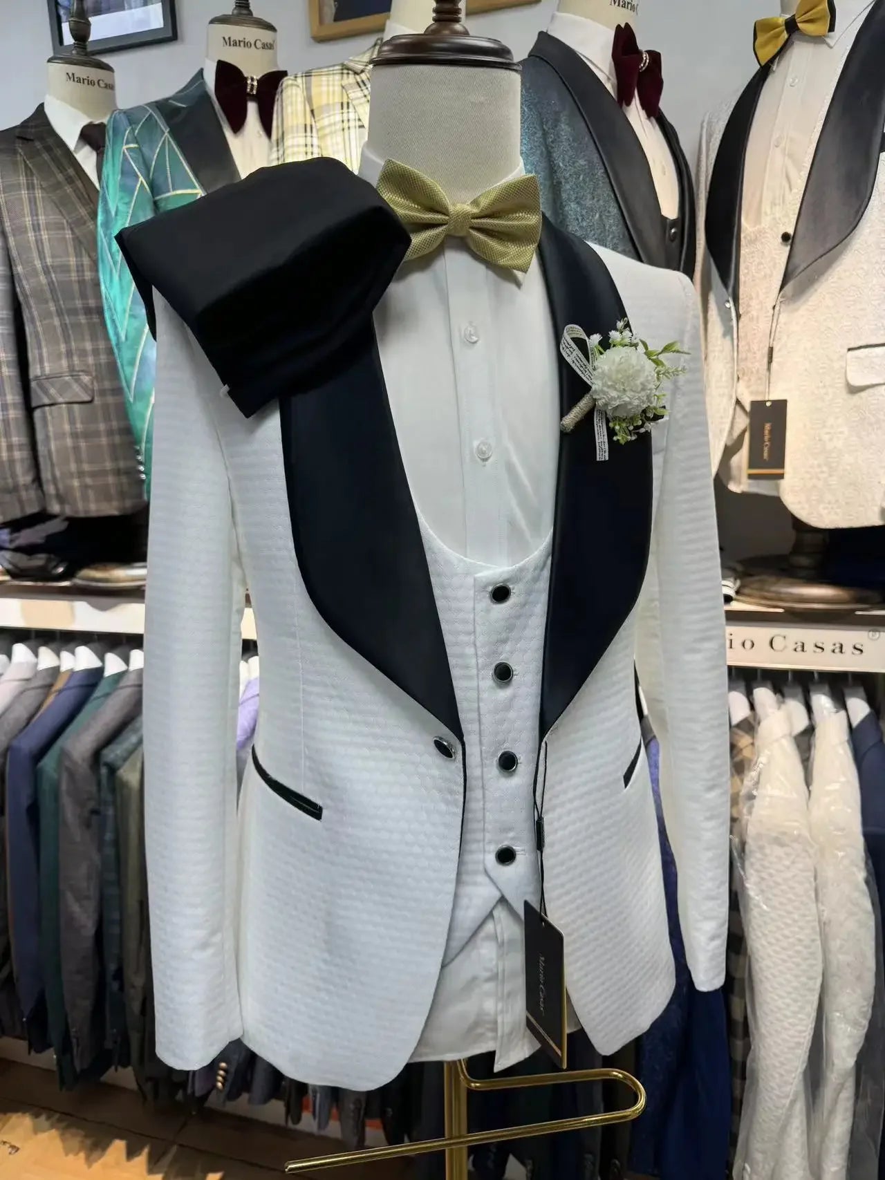Mannequin wearing a white tuxedo with black lapels, a gold bow tie, and a boutonnière, displayed in a formal wear store with various jackets in the background.