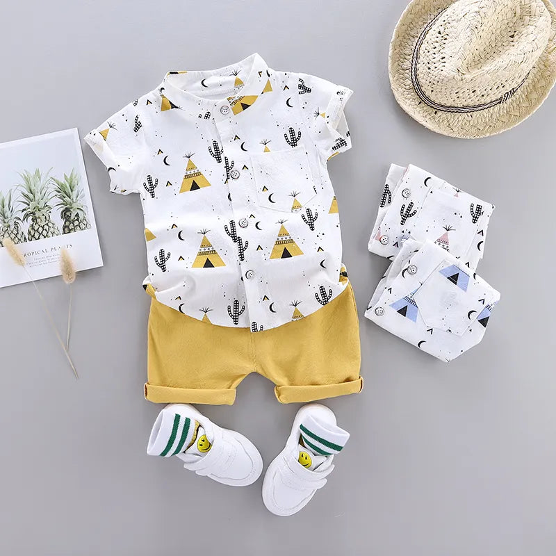 Children’s outfit with a white short-sleeve shirt featuring teepee and cactus prints, yellow shorts, white shoes with smiley faces, a straw hat, and a tropical-themed postcard.