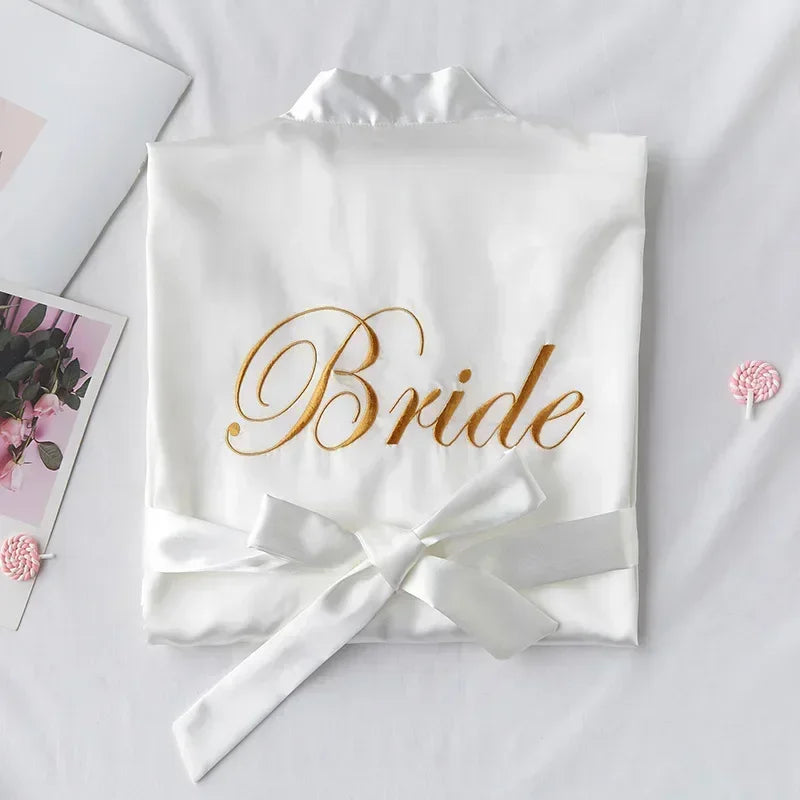 White silk robe with "Bride" embroidered in gold, neatly folded with a satin bow on a bed. Nearby are a pink floral card and a pink lollipop.