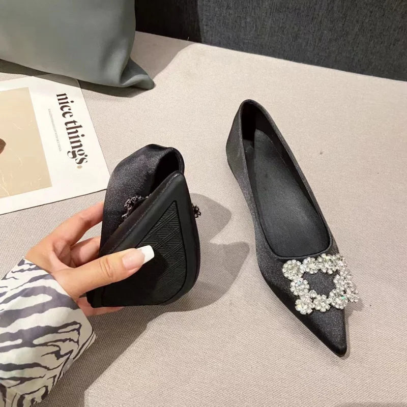 A hand holding a flexible, foldable black flat shoe with a textured sole. Another identical shoe with a decorative rhinestone buckle is placed on a light-colored surface nearby.