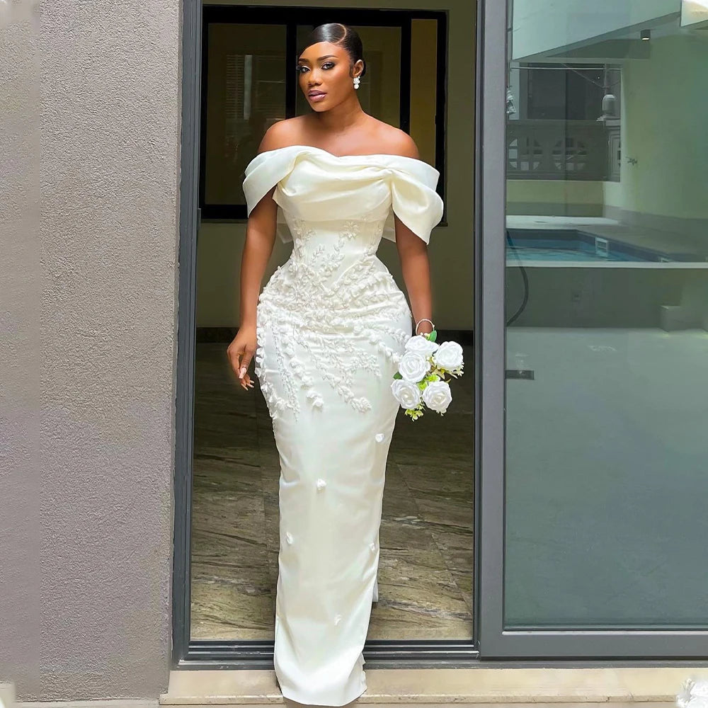 Woman in an elegant, off-shoulder white gown adorned with floral details, holding a bouquet of white roses, stands in a doorway with her hair styled up and wearing earrings.