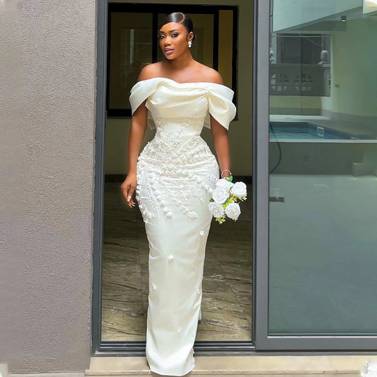 Woman in an elegant, off-shoulder white gown adorned with floral details, holding a bouquet of white roses, stands in a doorway with her hair styled up and wearing earrings.