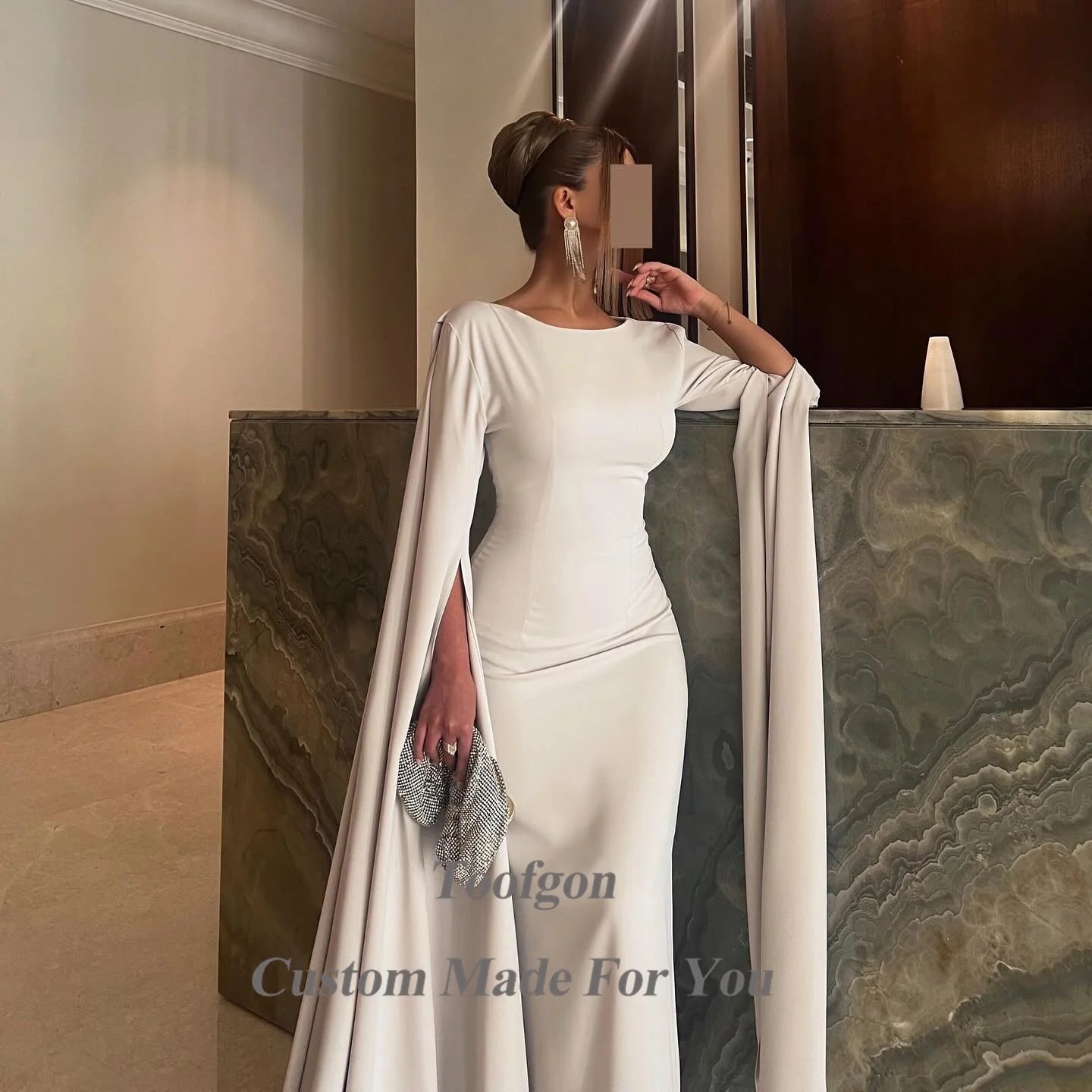 Elegant woman in a flowing white gown with cape sleeves, holding a sparkling clutch. She stands by a textured wall, wearing long silver earrings and an updo hairstyle.