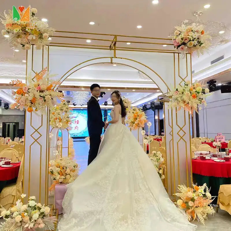 A bride and groom stand under an ornate floral arch in a decorated hall. The bride wears a large, elegant white gown, and the groom is in a black suit. Tables are set for a celebration.
