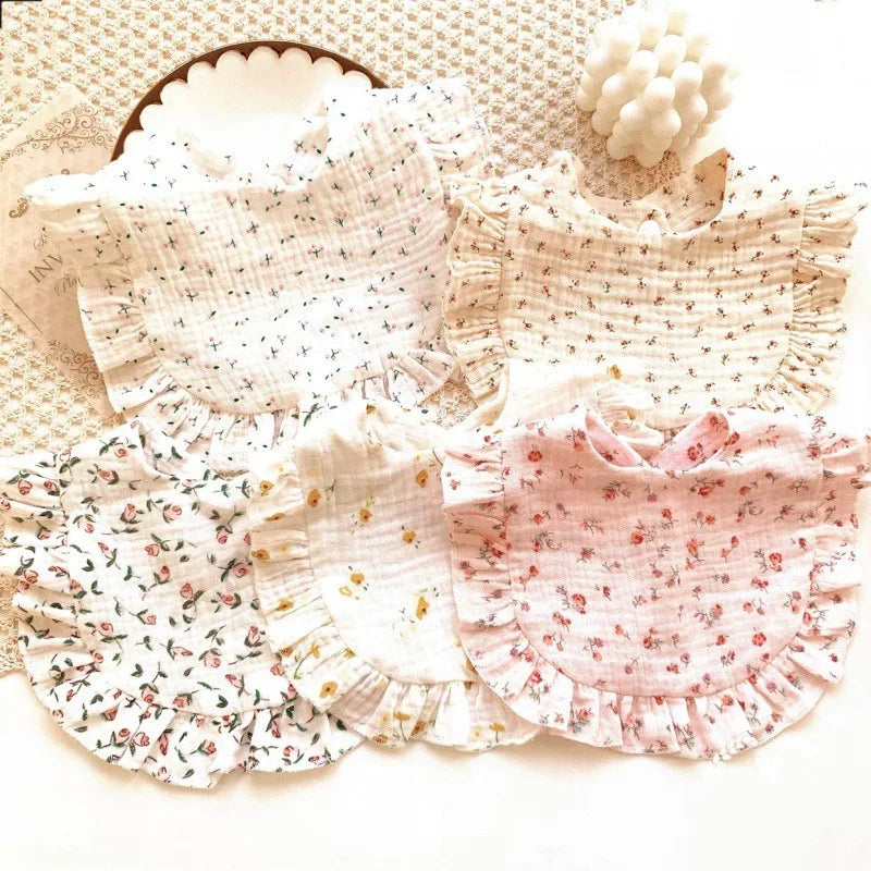 Five baby bibs with various floral patterns and ruffled edges are displayed on a textured white surface, alongside a decorative candle in the shape of small spheres.