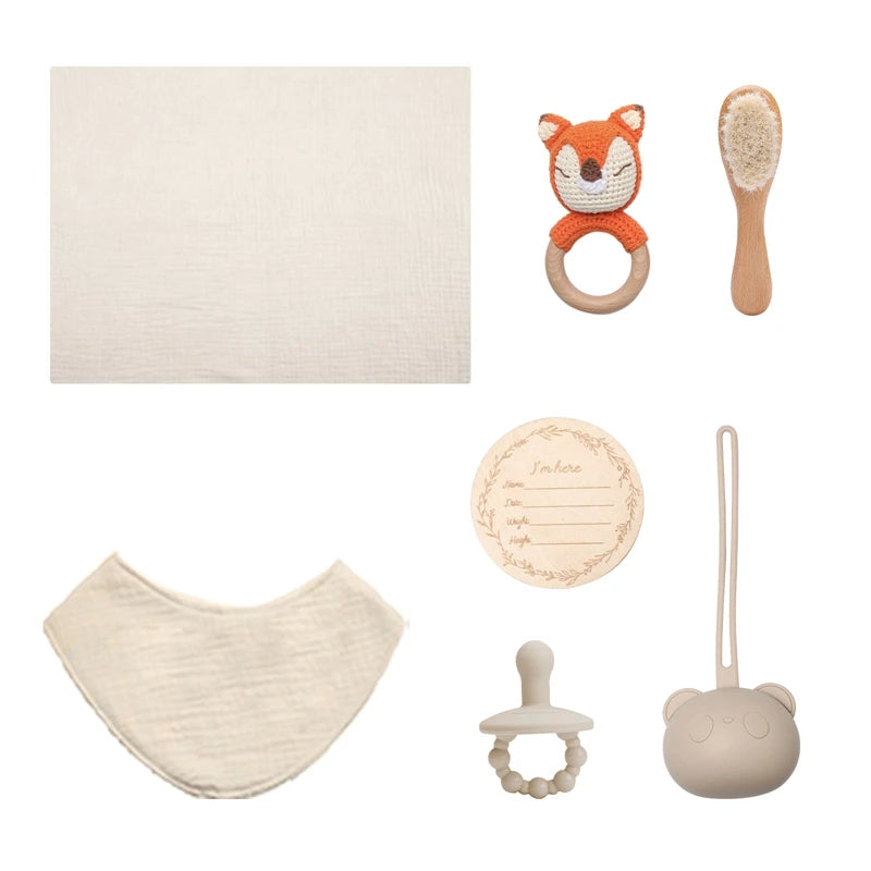 A baby gift set featuring a fox rattle, soft brush, pacifier with beads, bib, blanket, silicone case, and a wooden birth announcement disc. All items are in neutral tones.