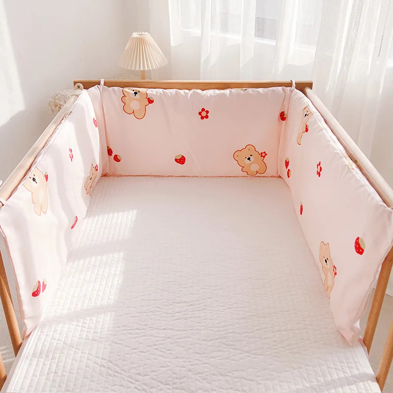 Crib with light pink padded bumpers featuring teddy bear and strawberry designs, set against a sunlit room backdrop with sheer curtains and a small lamp.