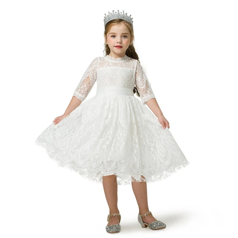 A young girl in a white lace dress and sparkling crown stands confidently, holding the dress' sides. She wears glittery shoes and has a red braided hairstyle, set against a white background.