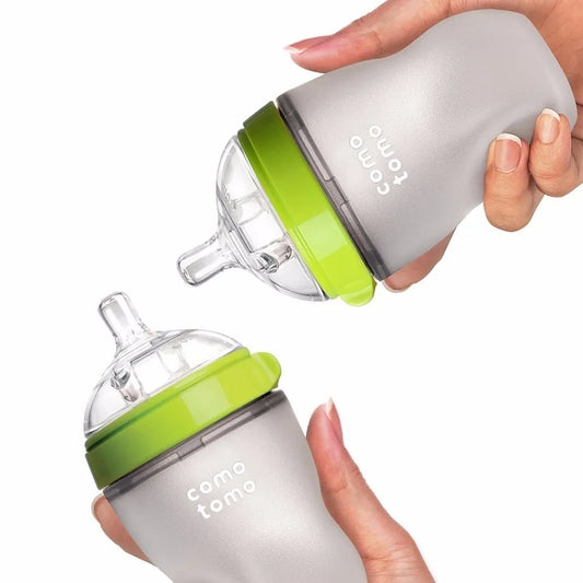 Two baby bottles with green lids and clear silicone nipples. One bottle is held upright, while the other is angled. The brand name is visible on the gray surface.