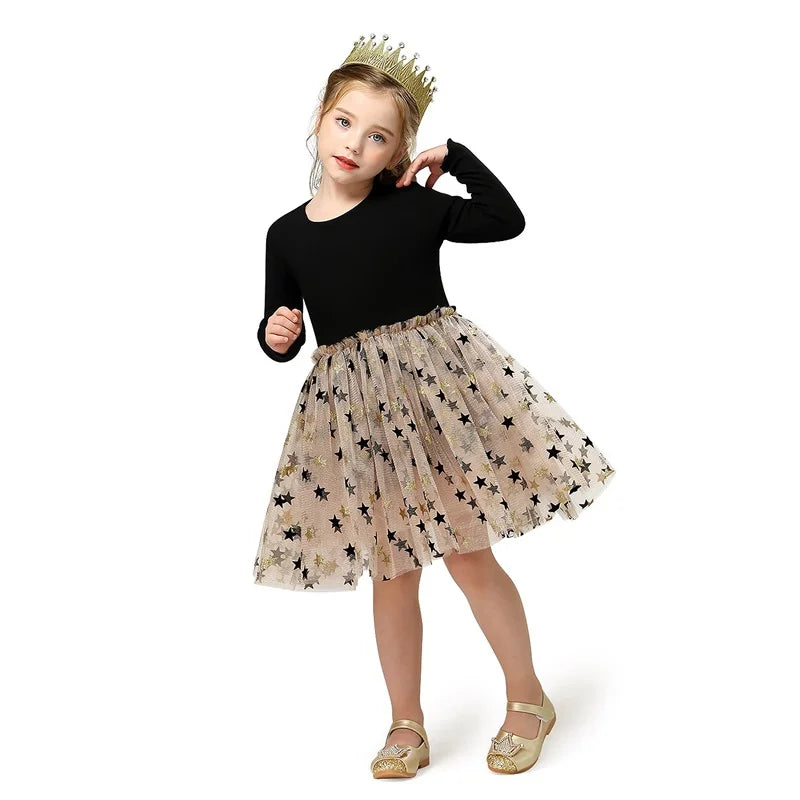A young girl wearing a gold crown, black top, and star-patterned tulle skirt poses against a white background. She has gold shoes and strikes a playful, confident pose.