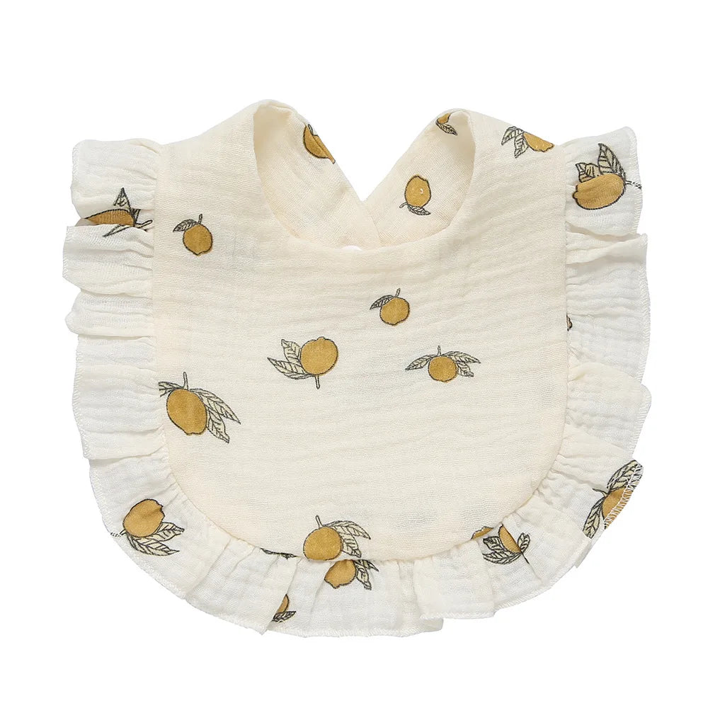 Cream-colored baby bib with ruffled edges, featuring a yellow lemon pattern with green leaves.