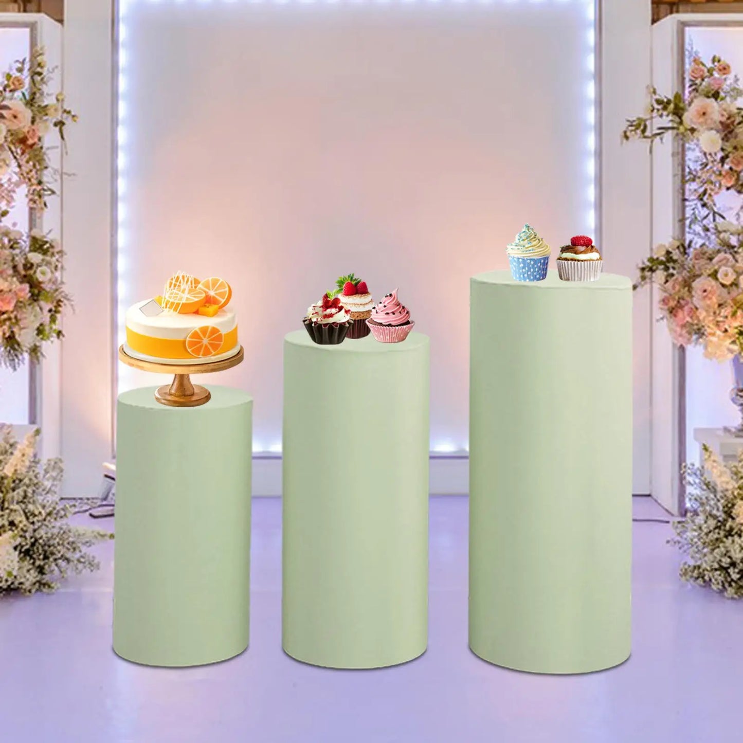 Three pastel green pedestals display desserts: a citrus cake, assorted cupcakes, and a blue polka dot cupcake. Background features floral arrangements and soft lighting.
