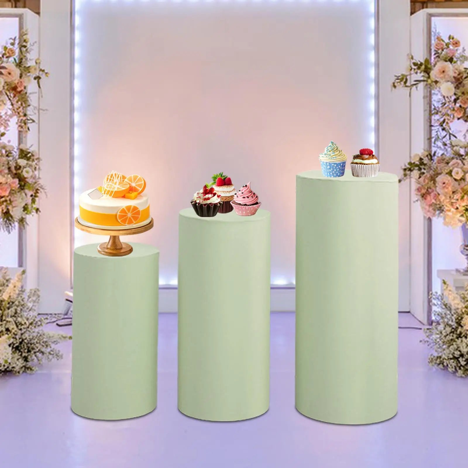 Three pastel green pedestals display desserts: a citrus cake, assorted cupcakes, and a blue polka dot cupcake. Background features floral arrangements and soft lighting.