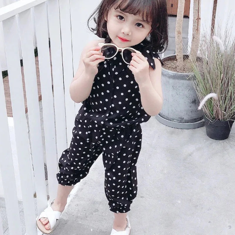 Young child in a black polka dot outfit, holding sunglasses, stands on a patio with potted plants, near a white fence. She wears white sandals with a playful expression.