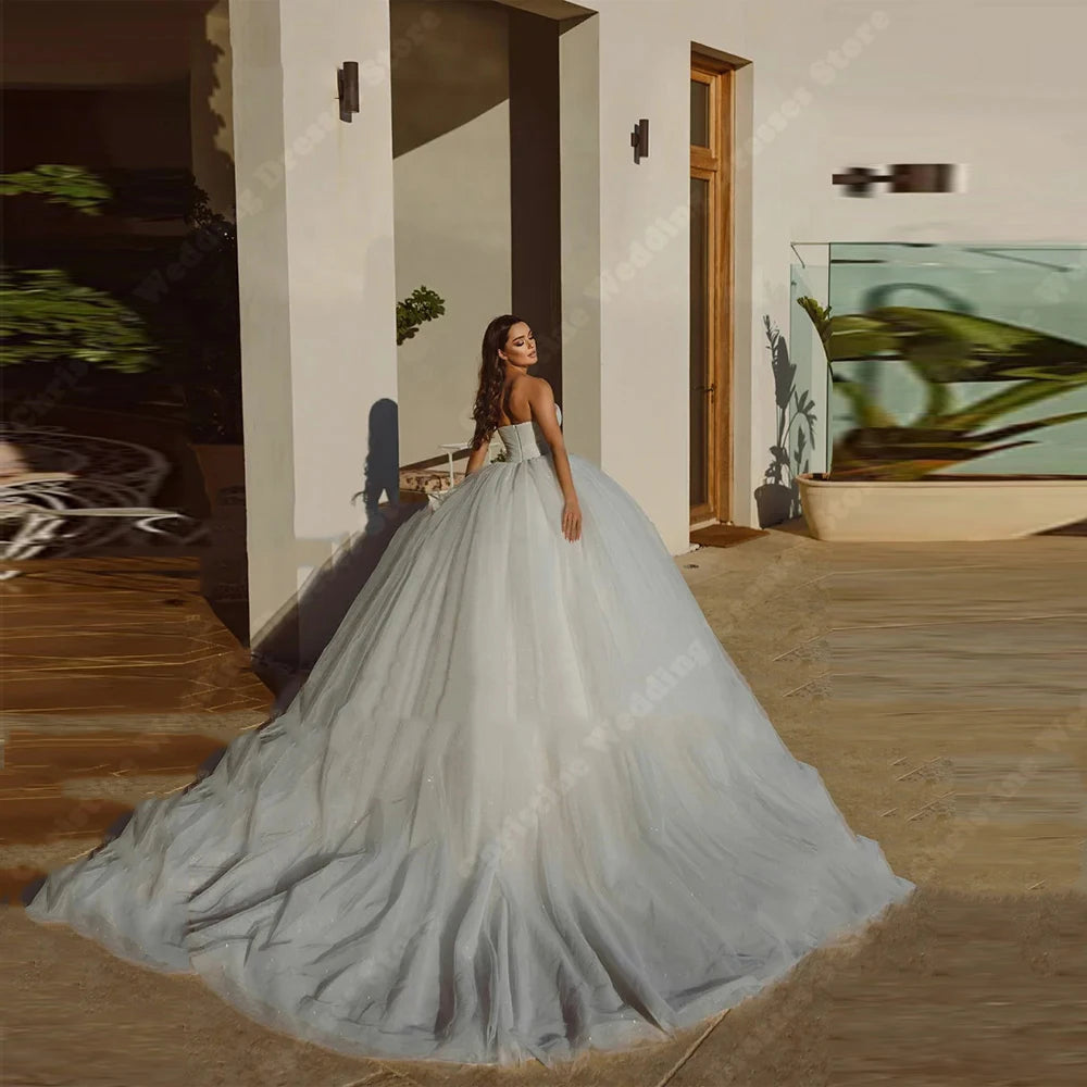 Woman in a strapless ball gown with a long, flowing train stands outdoors near a modern building, with plants and shadow play creating an elegant atmosphere.