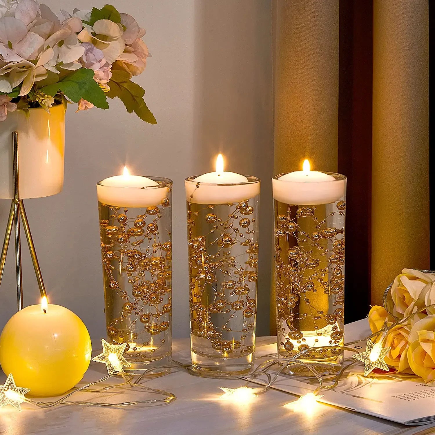 Three lit candles in tall glasses adorned with golden beads, surrounded by floral decor and star-shaped string lights, creating a warm and elegant ambiance.