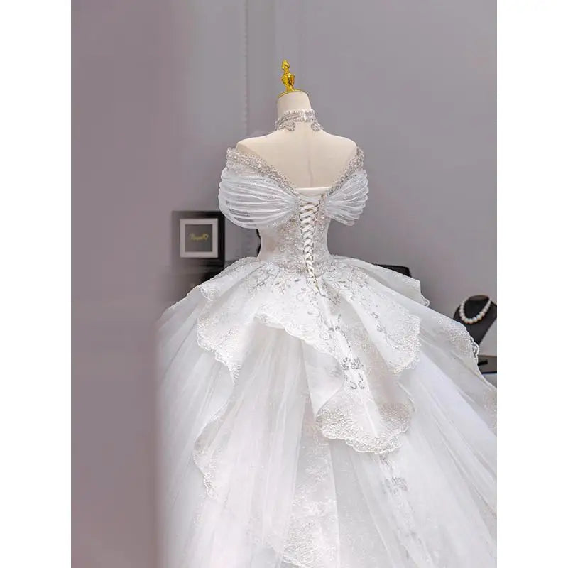 Elegant white wedding gown with an open back and lace-up detail, featuring off-shoulder tulle sleeves and layered lace skirt on a mannequin in a stylish room.
