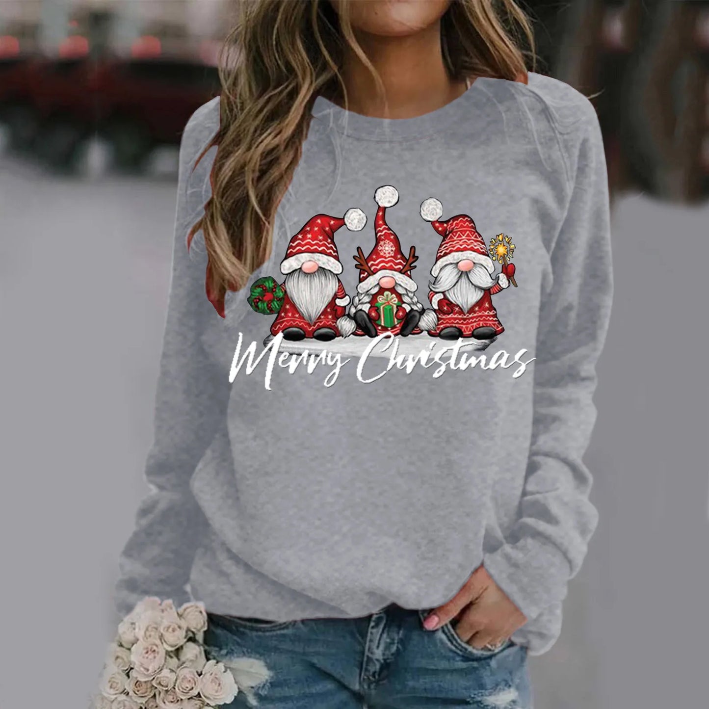 Woman wearing a gray sweater with festive gnome illustrations and "Merry Christmas" text. She holds white roses and is dressed in blue jeans. Background is blurred.