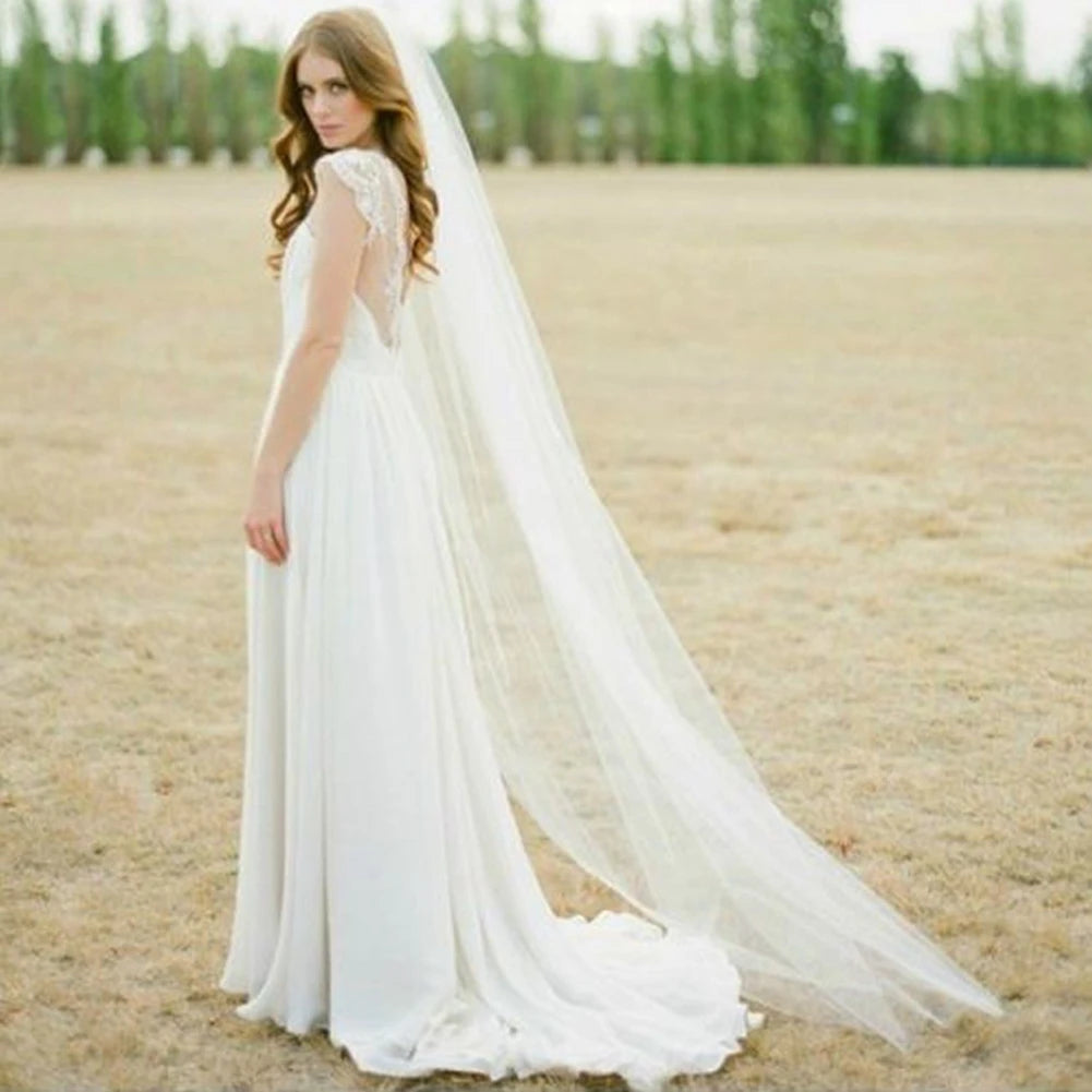 A bride in a flowing white wedding dress and long veil stands in a dry, open field with trees in the background. She's looking over her shoulder, her long hair cascading down.