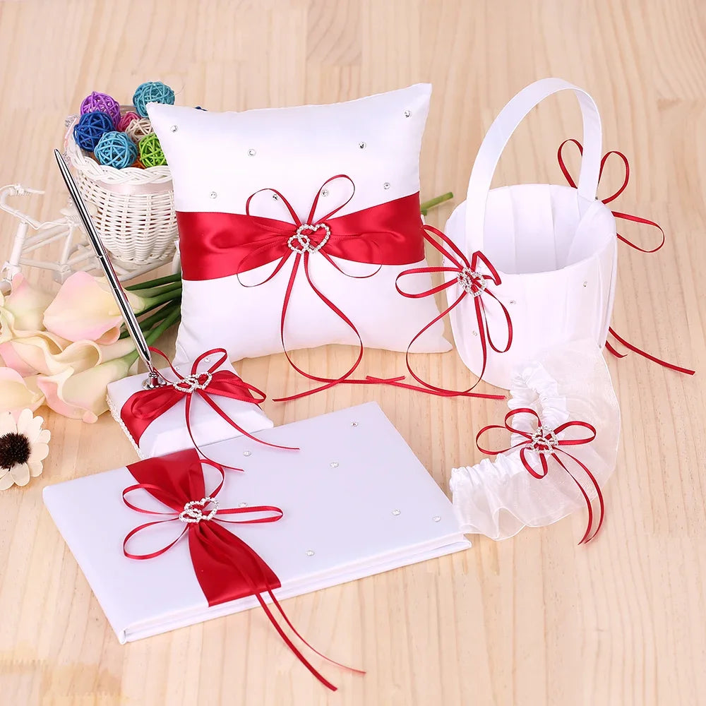 Wedding accessories set with red satin ribbons and rhinestones, including a pillow, basket, guest book, pen, and garter on a wooden table with flowers and colorful decorative balls.