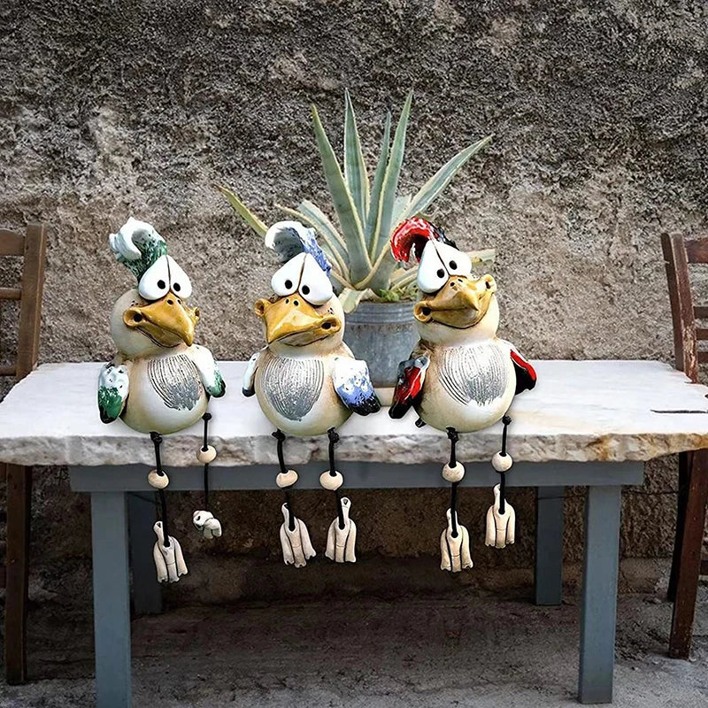 Three whimsical bird sculptures with quirky expressions sit on a stone table. Each bird features unique painted details in green, blue, and red. A potted plant is in the background.