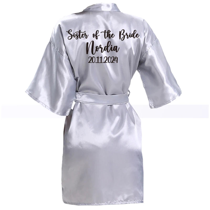 Silver satin robe with "Sister of the Bride," "Nordia," and "20.11.2024" in black script on the back. The robe is belted and has a glossy finish.