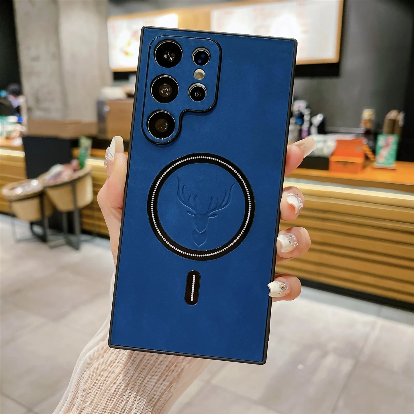A hand holding a blue smartphone with a deer emblem and multiple camera lenses on the back. The phone is in focus with a blurred café background featuring wooden accents.