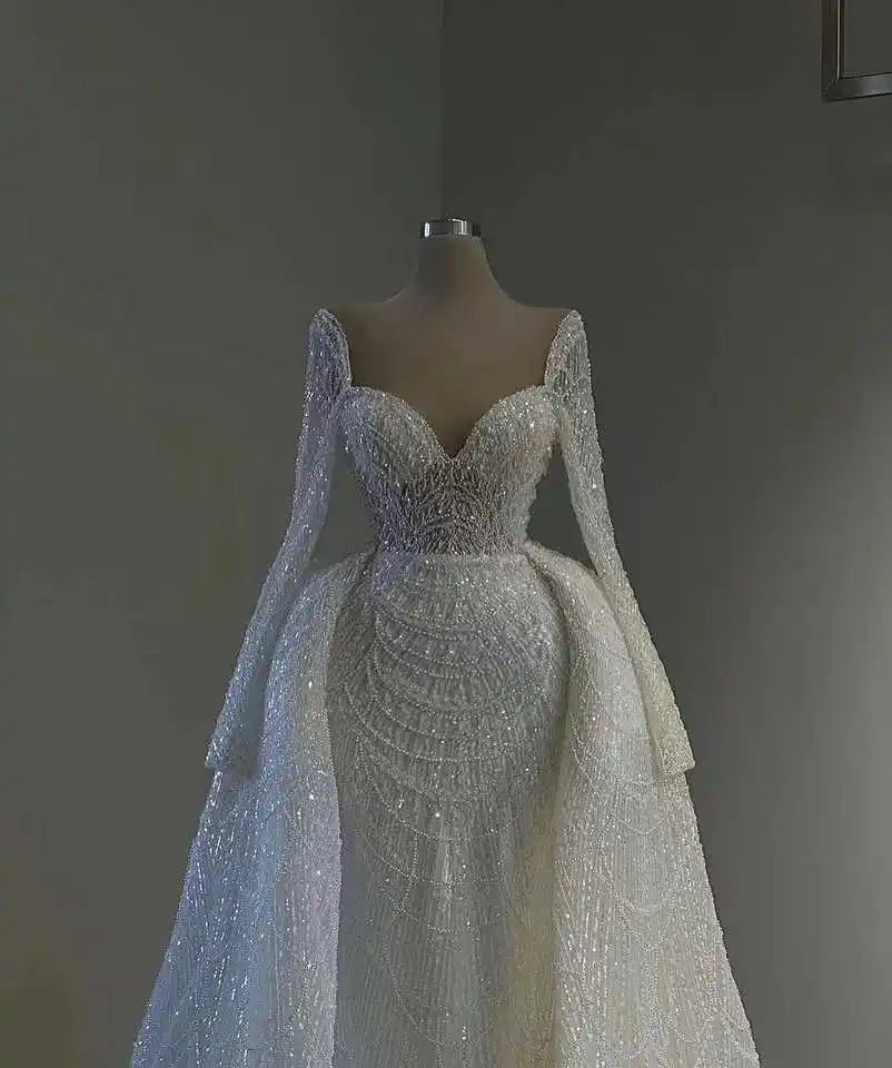 Elegant sparkling white wedding gown with intricate lace embroidery, sweetheart neckline, and long sheer sleeves on a mannequin against a neutral background.