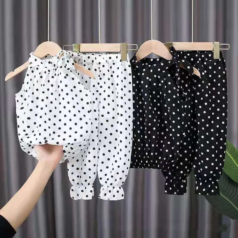 Three polka dot outfits hang on wooden hangers; two are black with white dots and one is white with black dots. A hand holds another white polka dot outfit.