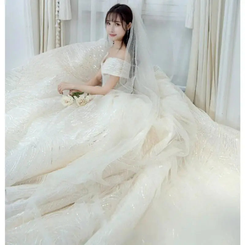 Bride in an elegant off-shoulder gown with sparkling details, seated and holding roses. She wears a veil, surrounded by flowing white fabric, gazing softly at the camera.